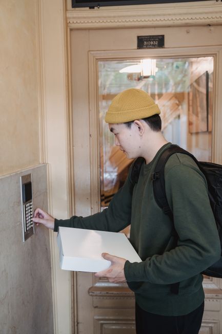 Asian male courier in beanie delivers package, pressing doorbell indoors.